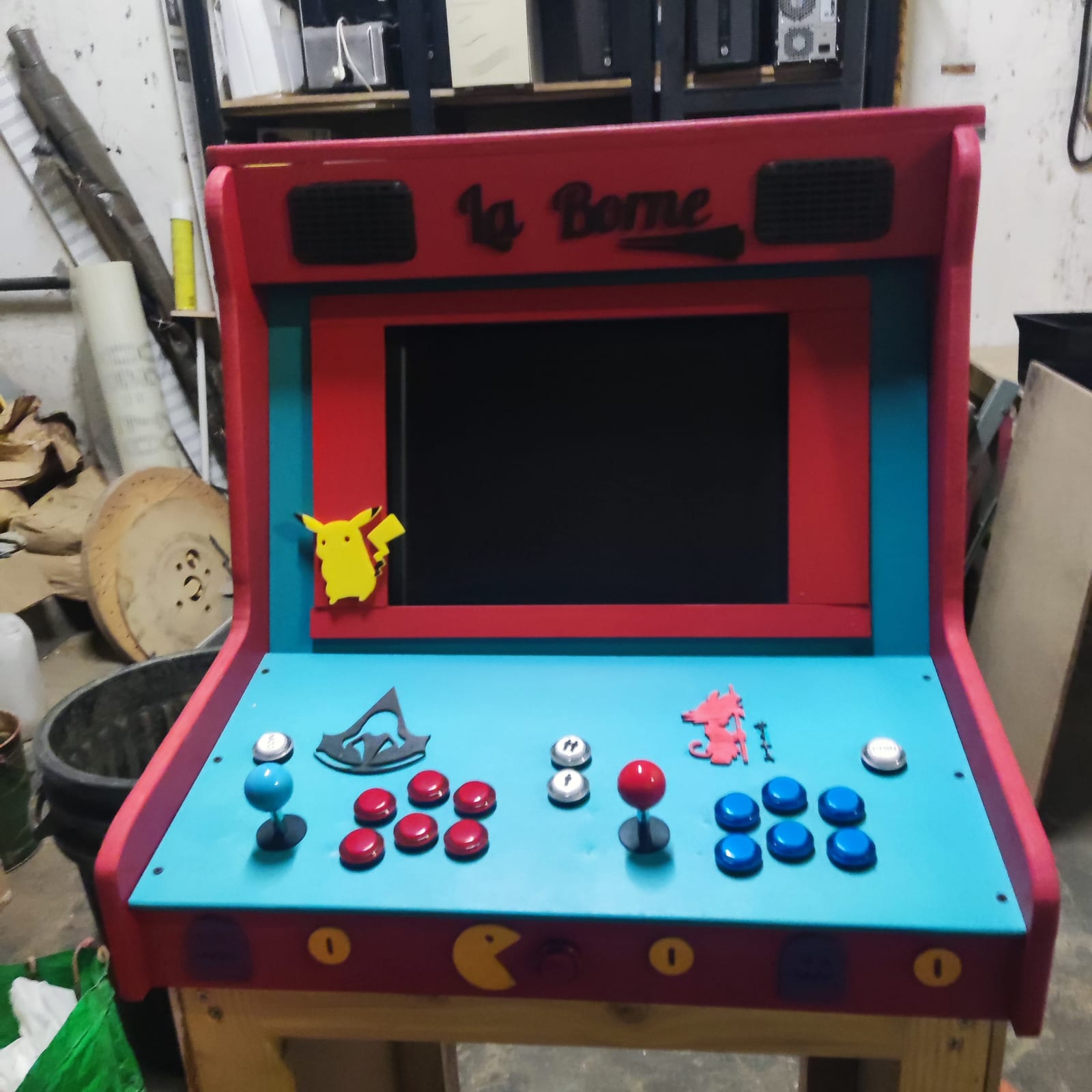La Borne terminée dans l'atelier : un caisson rouge et bleu posé sur un piétement de bois, son nom découpé sur l'enseigne entre deux grilles de haut-parleur, l'écran dans un cadre rouge, et le panneau de commande bleu avec deux manches et leurs boutons