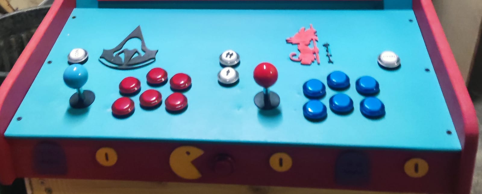 Le panneau de commande bleu vu de près : à gauche un manche à boule bleue et six boutons rouges, à droite un manche à boule rouge et six boutons bleus, des boutons argentés aux deux bouts et au milieu, et sur la façade rouge un Pac-Man et des pièces peintes