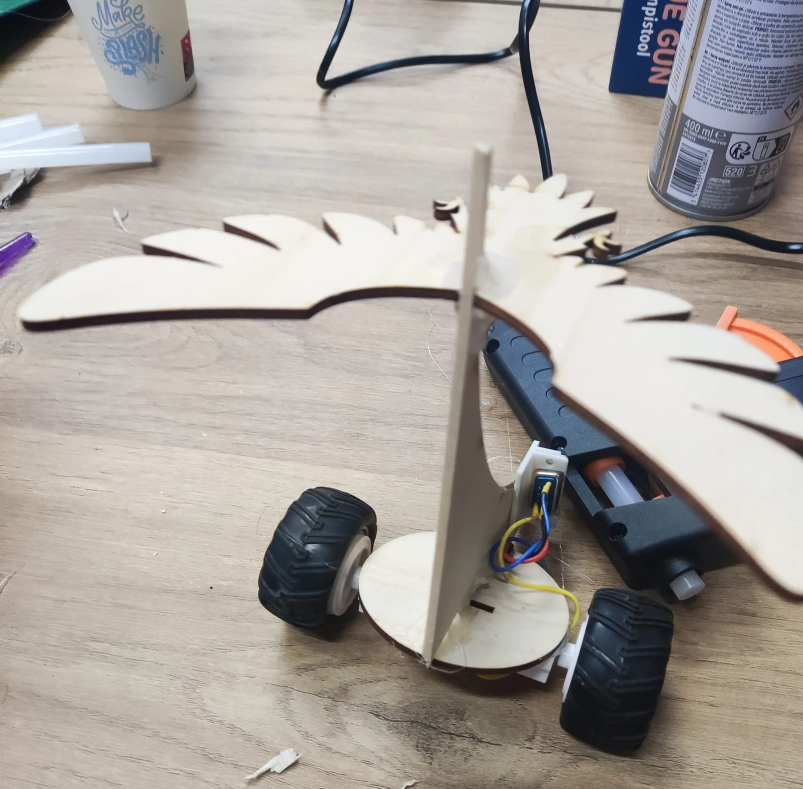 Un robot en cours de montage sur l'établi : un oiseau de contreplaqué découpé au laser, planté sur un socle rond à deux grosses roues, ses moteurs raccordés par un domino, le pistolet à colle posé derrière