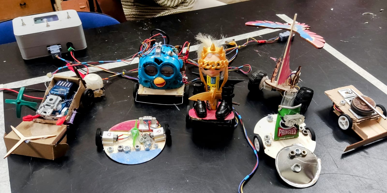Sept robots bricolés alignés sur une table noire, reliés par leurs câbles à un boîtier de commande gris : un Furby, une poupée couronnée, un oiseau de contreplaqué, une boîte de Pringles, des socles de carton et de bois sur des roues