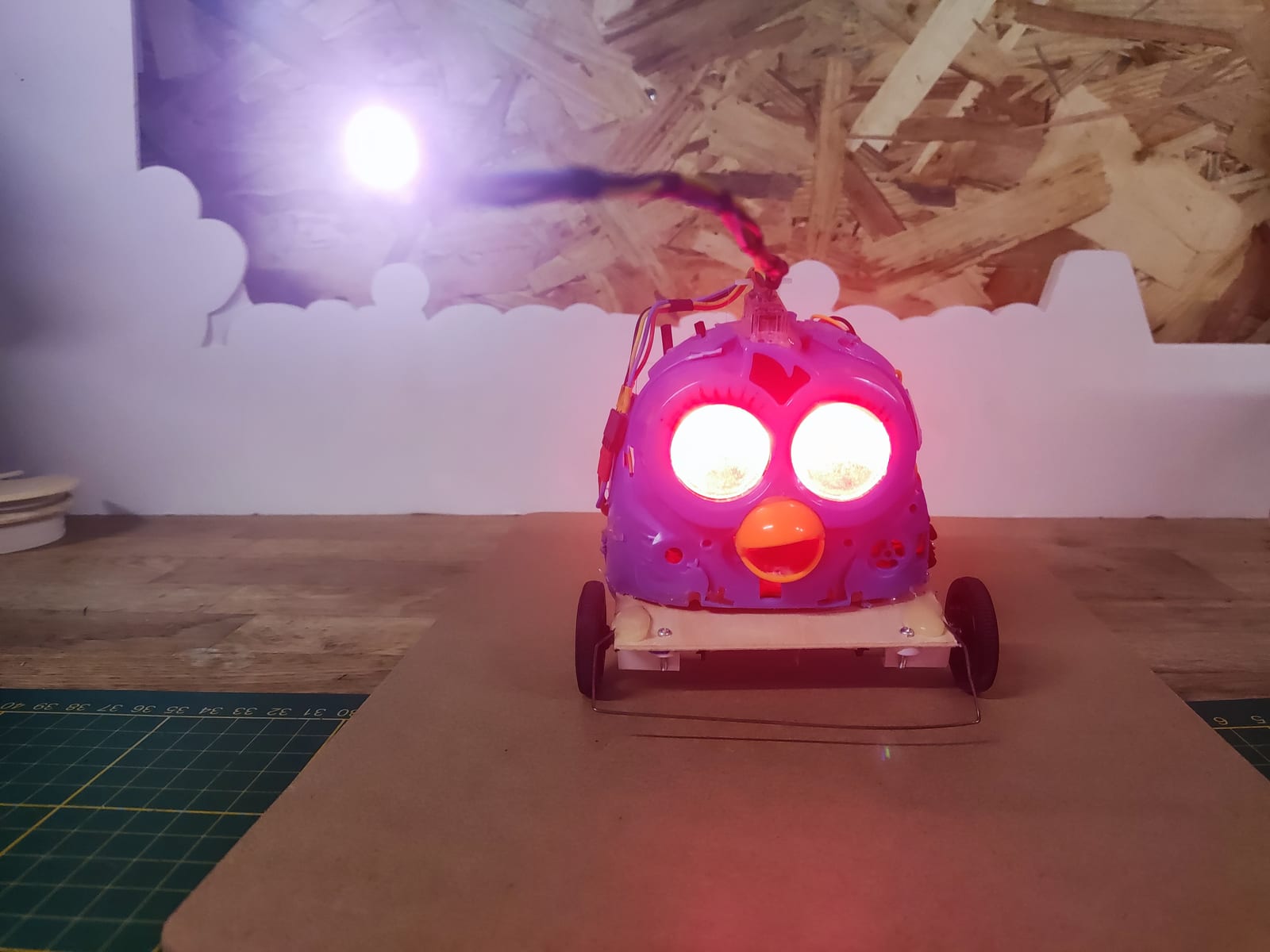 Le Furby-Bricobot en janvier 2026, les yeux allumés en rose et une lampe violette au bout d'une tige, posé sur une planche devant un décor aux créneaux blancs