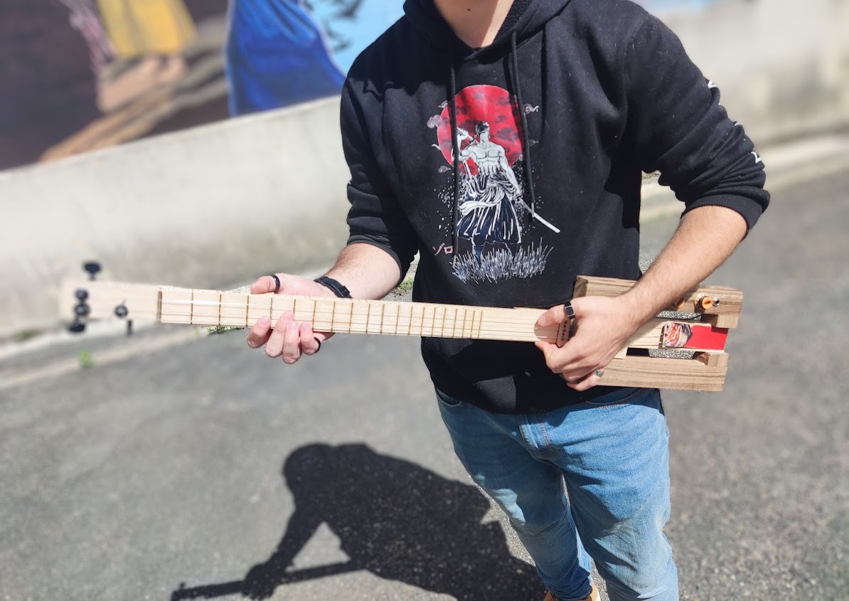 Une guitare cigar box tenue à deux mains devant un mur peint : un long manche clair garni de frettes, trois mécaniques sur la tête, et au bout une boîte en bois ouverte sur le côté