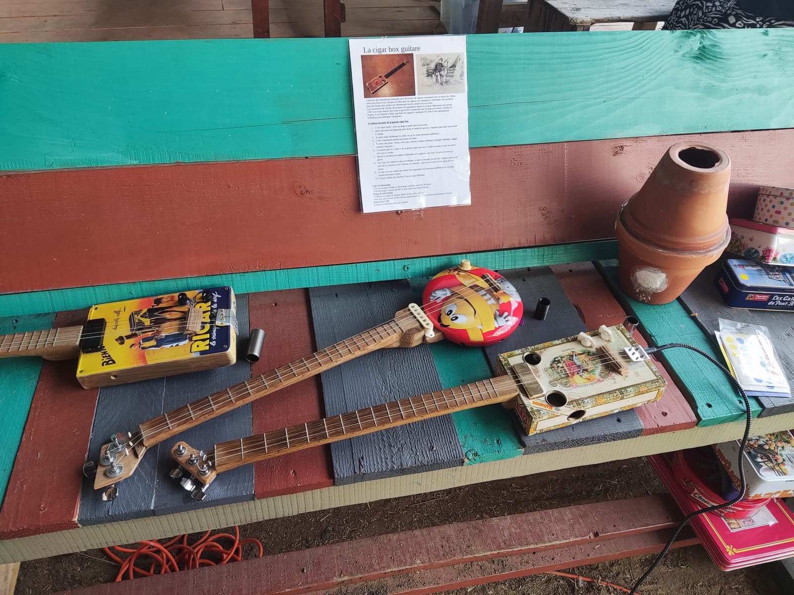 Trois guitares cigar box posées sur un banc de planches peintes : l'une sur une boîte en fer jaune, l'autre sur une boîte en fer ronde, la troisième sur une boîte à cigares en bois reliée à un câble, et au-dessus l'affichette de l'atelier