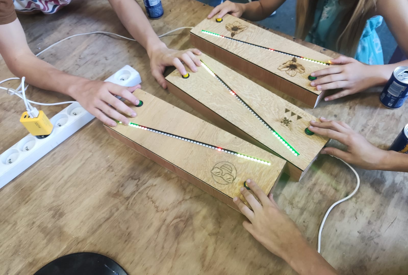 Trois Pong-LED réduits posés en éventail sur un établi, des mains sur les boutons à chaque bout, les bandes allumées en biais sur des couvercles gravés de fleurs et de motifs